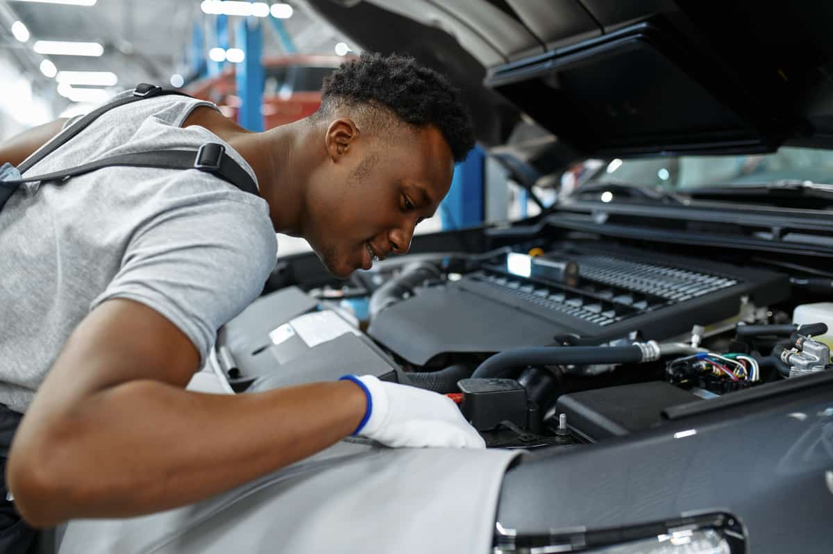 Male mechanic checks engine, car service. Vehicle repairing garage, man in uniform, automobile station interior on background
